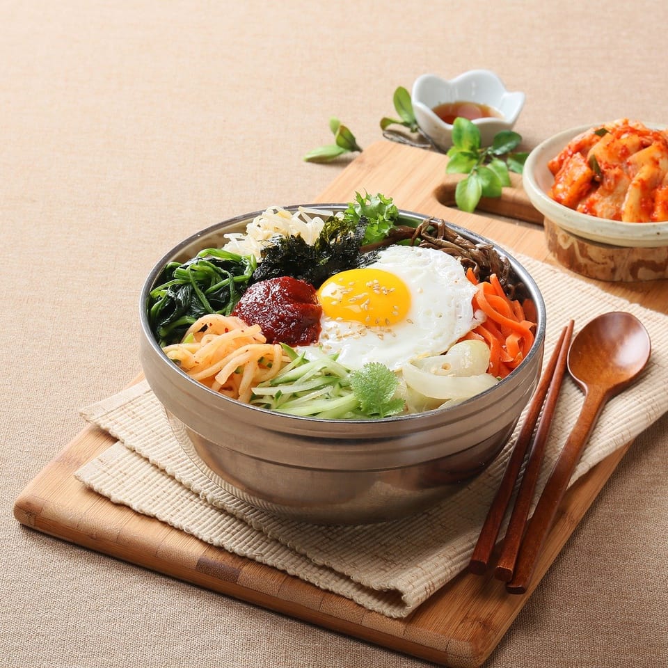 Korean bibimbap topped with a fried egg, vegetables, seaweed, and red pepper paste, served with kimchi and utensils.