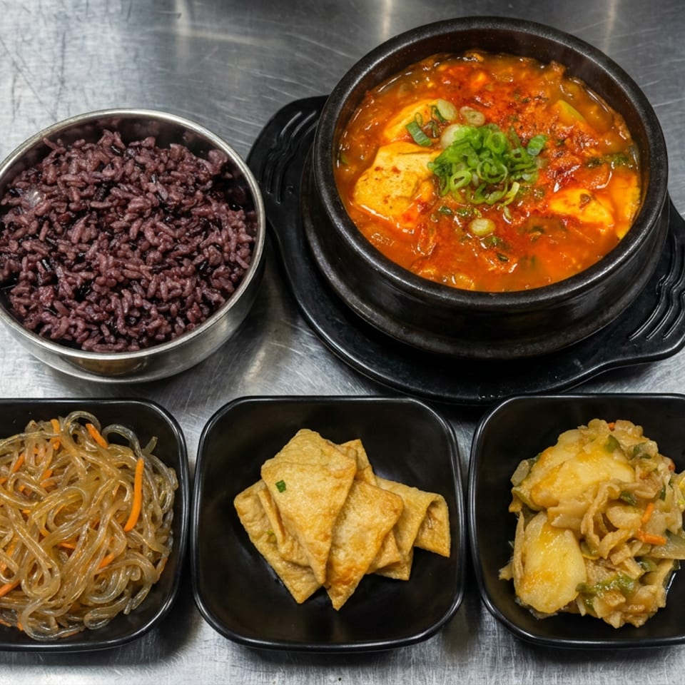Korean meal with purple rice, spicy tofu stew, glass noodles, fried tofu, and vegetable fritters on a metal table.