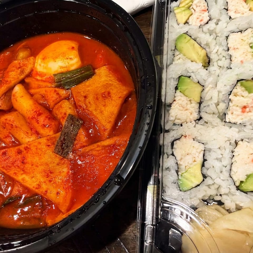Spicy Korean rice cakes and fish cakes sit beside avocado sushi rolls with creamy seafood topping.